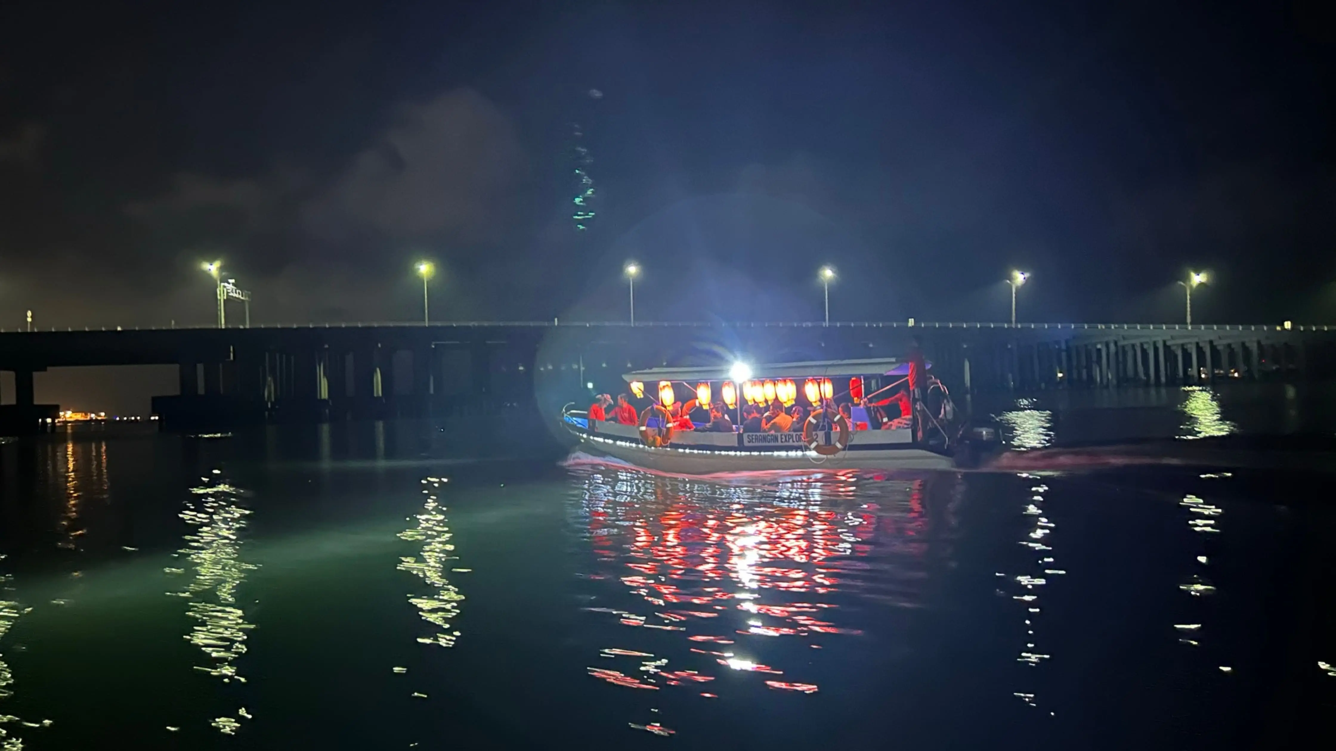 A mangrove night cruise boat in Bali illuminated with lanterns, sailing under the bridge lights on calm evening waters